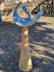 Eine Sonnenuhr aus Metall mit einer skulpturalen Form steht auf einem gepflasterten Platz. Der Hintergrund zeigt eine Steinmauer und eine charakteristische Umgebung.