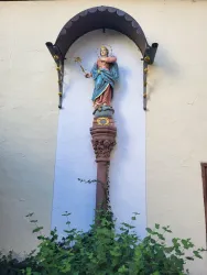 Eine Statue von Maria steht auf einer Säule vor einer Wand. Um die Statue herum wachsen grüne Pflanzen.