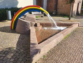 Ein schöner Brunnen mit einem bunten Regenbogen aus Rohren. Wasser sprudelt aus der Skulptur und fließt in die flache Steinform.