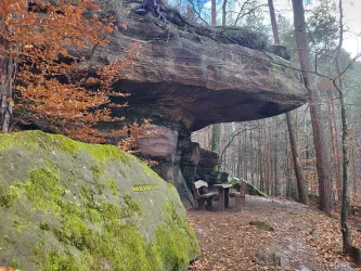 Eine beeindruckende Felsformation mit einer Überhänge und einer Holzbank darunter. Im Hintergrund sind Bäume und herbstliches Laub zu sehen.