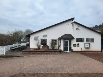 Ein modernes Gebäude mit einem hellen Außenanstrich und einem schrägen Dach. Vor dem Gebäude befindet sich ein gepflasterter Bereich mit Sitzgelegenheiten und Pflanzen.