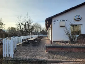 Ein ruhiger Außenbereich mit Bänken und einem weißen Zaun. Im Hintergrund ist ein Gebäude mit einem Schild sichtbar.