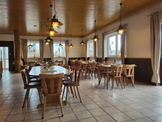 Ein gemütlicher Speisesaal mit Holztischen und Stühlen. Die Beleuchtung ist warm und einladend, mit Fenstern, die Tageslicht hereinlassen.