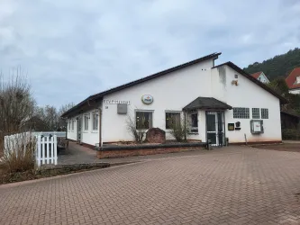 Ein modernes Gebäude mit einem Eingang und Fenstern. Der Parkplatz vor dem Gebäude ist gepflastert und es gibt einige Pflanzen in der Umgebung.