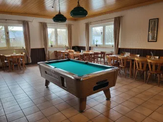 Ein gemütlicher Raum mit einem Billardtisch in der Mitte. Rundherum stehen Holztische und Stühle, und große Fenster lassen viel Licht herein.