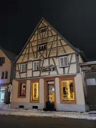 Ein traditionelles Fachwerkhaus mit einer hellen Fassade. Es ist nachts und die Fenster sind beleuchtet, während Schnee auf dem Boden liegt.