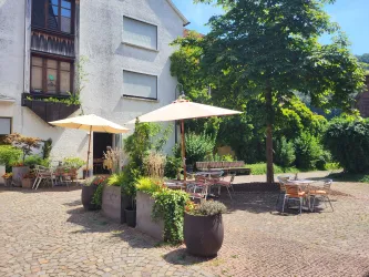 Ein gemütlicher Außenbereich mit Tischen und Sonnenschirmen. Umgeben von Pflanzen und einem ruhigen Ambiente.