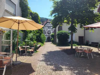 Eine idyllische Innenhof mit Bäumen und Sonnenschirmen. Es gibt gemütliche Sitzgelegenheiten und historische Gebäude im Hintergrund.