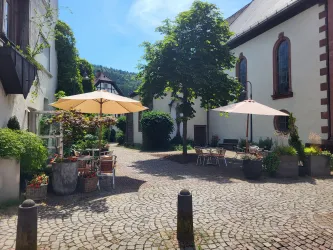 Ein malerischer Innenhof mit Steinen und Sonnenschirmen. Üppiges Grün und gemütliche Sitzgelegenheiten laden zum Verweilen ein.