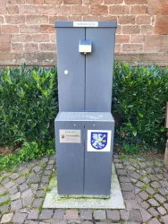Ein grauer Trinkbrunnen steht auf einem gepflasterten Weg umgeben von grünen Pflanzen.