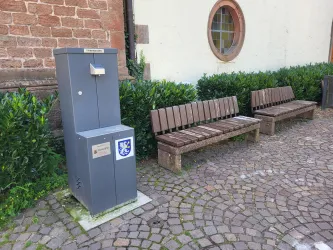 Ein grauer Wasserautomat steht neben zwei Holzbänken. Die Umgebung besteht aus gepflastertem Boden und üppigem Grün.