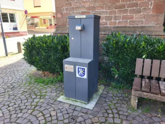 Ein grauer Trinkbrunnen steht auf einem gepflasterten Platz. Er ist von grünen Pflanzen umgeben und in der Nähe befindet sich eine Bank.