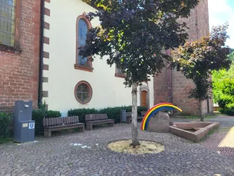 Ein ruhiger Platz mit Bänken und einem Baum vor einer alten Kirche. Im Hintergrund ist ein farbenfrober Regenbogen dekorativ aufgestellt.