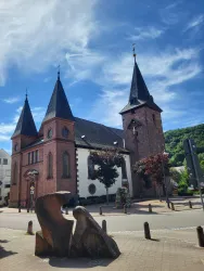 Eine charmante Kirche mit zwei Türmen und einer attraktiven Fassade aus roten Ziegeln. Im Vordergrund steht eine moderne Skulptur, umgeben von einer malerischen Landschaft.