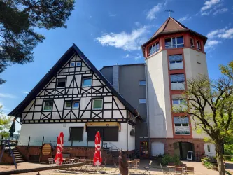 Ein traditionelles Fachwerkhaus mit einem modernen Turm-Anbau. Im Vordergrund sind Terrassenmöbel und Sonnenschirme zu sehen.