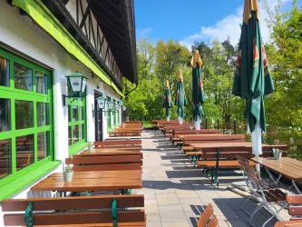 Ein Außenbereich eines Restaurants mit Holzstühlen und -tischen. Im Hintergrund sind grüne Bäume und Sonnenschirme zu sehen.