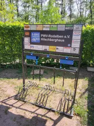 Eine Fahrradabstellanlage mit mehreren Abstellplätzen und einem Schild des PVW-Rodalben e.V. Es gibt Informationen zu E-Bikes und Sponsoren auf dem Schild.