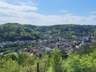 Eine malerische Landschaft mit einer kleinen Stadt, umgeben von grünen Hügeln und Bäumen. Der Himmel ist klar und sonnig.