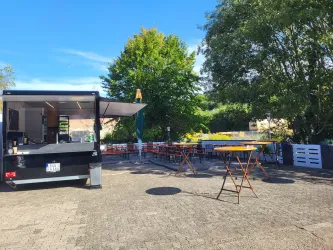 Ein Imbisswagen steht auf einem gepflasterten Platz mit einigen Tischen und Stühlen. Im Hintergrund sind grüne Bäume und ein blauer Himmel zu sehen.
