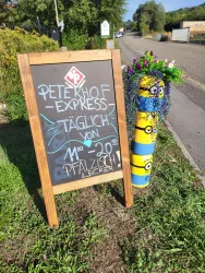 Ein Schild des Peterhof Express mit einem bunten Design steht am Wegesrand. Es informiert über die täglichen Öffnungszeiten und bietet pfälzische Spezialitäten an.