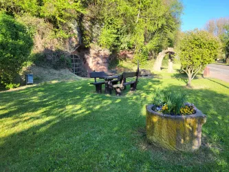Eine grüne Wiese mit einem Tisch und Bänken zum Sitzen. Im Hintergrund sind Bäume und ein kleiner Steinbrunnen mit Blumen zu sehen.