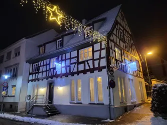 Ein schön beleuchtetes Fachwerkhaus bei Nacht, geschmückt mit Lichtern und einem Stern. Auf dem Boden liegt etwas Schnee.