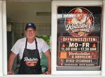 Ein freundlich lächelnder Mann steht am Fenster eines Imbisses. Im Hintergrund ist ein Schild mit den Öffnungszeiten und Informationen zu sehen.