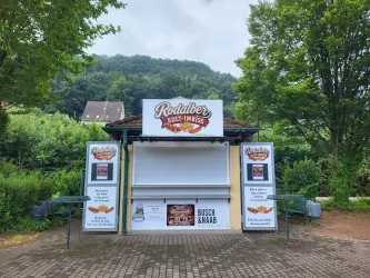 Ein Imbissstand namens Rodalber Kultimbiss steht in einer grünen Umgebung. Es gibt eine leere Verkaufstheke und informative Schilder auf beiden Seiten.