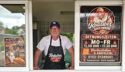 Ein freundlicher Imbissbetreiber steht an einem Fenster seines Standes. Im Hintergrund sind Werbetafeln mit Öffnungszeiten und Angeboten zu sehen.
