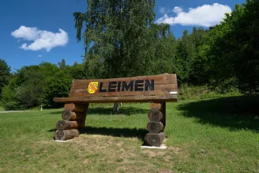 Ein Holzschild mit der Aufschrift "Leimen" steht in einer grünen Umgebung. Im Hintergrund sind Bäume und blauer Himmel zu sehen.