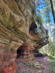 Eine beeindruckende Felsformation in einem Waldgebiet, umgeben von hohen Bäumen. Der Weg ist mit Laub bedeckt und es gibt - geschützt unter einem Felsvorsprung - Tische und Bänke zum Sitzen.