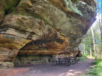 Eine beeindruckende Felsformation mit Überhang. Darunter befindet sich ein Tisch aus Holz und umgeben von Bäumen.