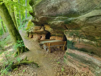 Eine natürliche Felsformation mit einem kleinen Tisch und Bänken in einem Wald. Der Weg führt sanft an der Felswand entlang, umgeben von Bäumen und Laub.