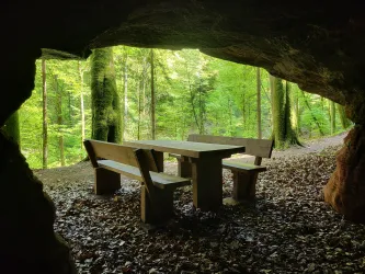 Ein Tisch mit Bänken in einer Höhle im Wald. Die Umgebung ist grün und von Bäumen umgeben.