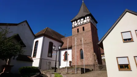 Ein historischer Turm mit einem spitzen Dach steht neben modernen Häusern. Der Himmel ist klar und blau.