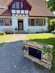 Ein charmantes Gebäude mit rot gedecktem Dach und weißen Wänden. Vor dem Eingang steht ein Holzschild mit Blumen und einer freundlichen Einladung.