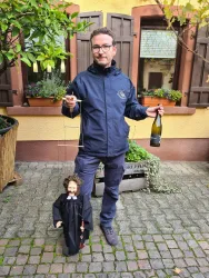 Ein Mann hält eine Flasche Sekt und eine Marionette in Form eines kleinen Mannes. Im Hintergrund sind Pflanzen und eine gemütliche Häuserwand zu sehen.