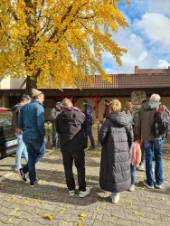 Eine Gruppe von Menschen steht auf einem gepflasterten Platz unter einem Baum mit gelben Blättern. Im Hintergrund ist ein Gebäude und ein blauer Himmel zu sehen.