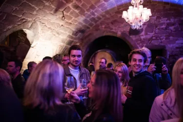 Eine lebhafte Veranstaltung in einem alten Gewölbe mit Gästen, die Drinks in der Hand halten. Im Hintergrund strahlt ein eleganter Kronleuchter Licht in den Raum.