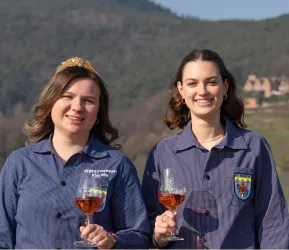 Zwei Frauen halten Gläser mit Wein und lächeln in die Kamera. Im Hintergrund sind sanfte Hügel und ein klarer Himmel zu sehen.