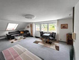 Ein helles Wohnzimmer mit einem Sofa, einem Tisch und Stühlen. Große Fenster bieten einen Blick ins Grüne.