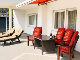 Eine sonnige Terrasse mit roten Stühlen und einem Tisch. Im Hintergrund sind Liegestühle und Fenster zu sehen.