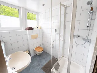 Ein modernes Badezimmer mit einer Dusche, einem Waschbecken und einer Toilette. Große Fenster sorgen für viel Tageslicht.