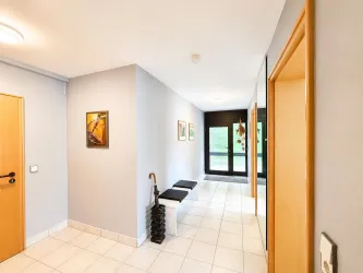 Ein heller Flur mit blaugrauen Wänden und weißen Fliesen. Auf der linken Seite befinden sich zwei Türen und Bilder an der Wand, während auf der linken Seite eine Bank zu sehen ist.