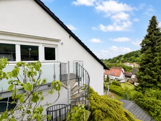 Ein modernes Haus mit einer geschwungenen Treppe und großen Fenstern. Im Hintergrund sind grüne Hügel und ein blauer Himmel zu sehen.