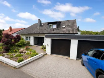 Ein modernes Haus mit einem begrünten Vorgarten und zwei Garagen. Ein blauer Wagen steht vor dem Eingang.