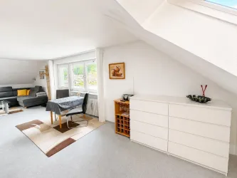 Helles Wohnzimmer mit einer gemütlichen Sitzecke und großen Fenstern. Ein Tisch mit Stühlen und eine moderne Kommode runden den Raum ab.