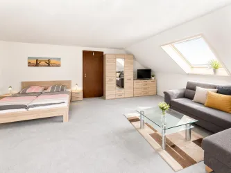 Ein helles Zimmer mit schrägen Fenstern, einem Doppelbett und einem komfortablen Sofa. Ein Glastisch und eine moderne Kommode vervollständigen die Einrichtung.