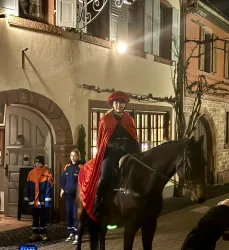 Eine Person in rot-grüner Kleidung sitzt auf einem Pferd und steht vor einem historischen Gebäude. Neben ihr steht ein Kind in Arbeitskleidung.