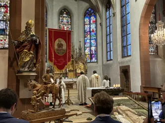 Ein religiöser Gottesdienst in einer alten Kirche mit bunten Fenstern. Im Vordergrund sind Statuen und eine Gruppe von Personen in liturgischer Kleidung zu sehen.